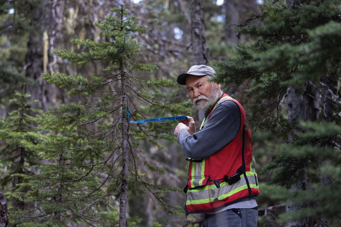 A photo of a bearded man in a baseball cap and orange work vest wraps a blue plastic trail marker around a tree.